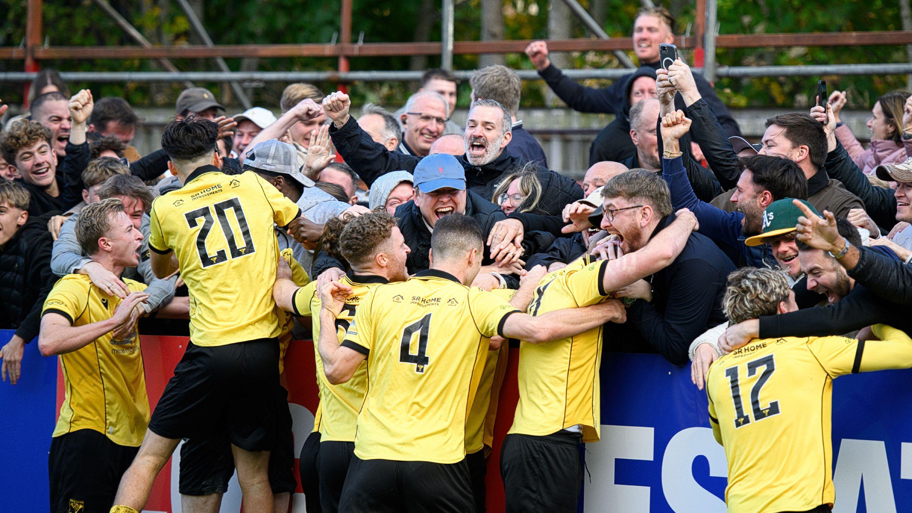 Football players in yellow kit celebrate with fans who are reaching out to them with expressions of triumph and happiness. Some are punching the air with fists.