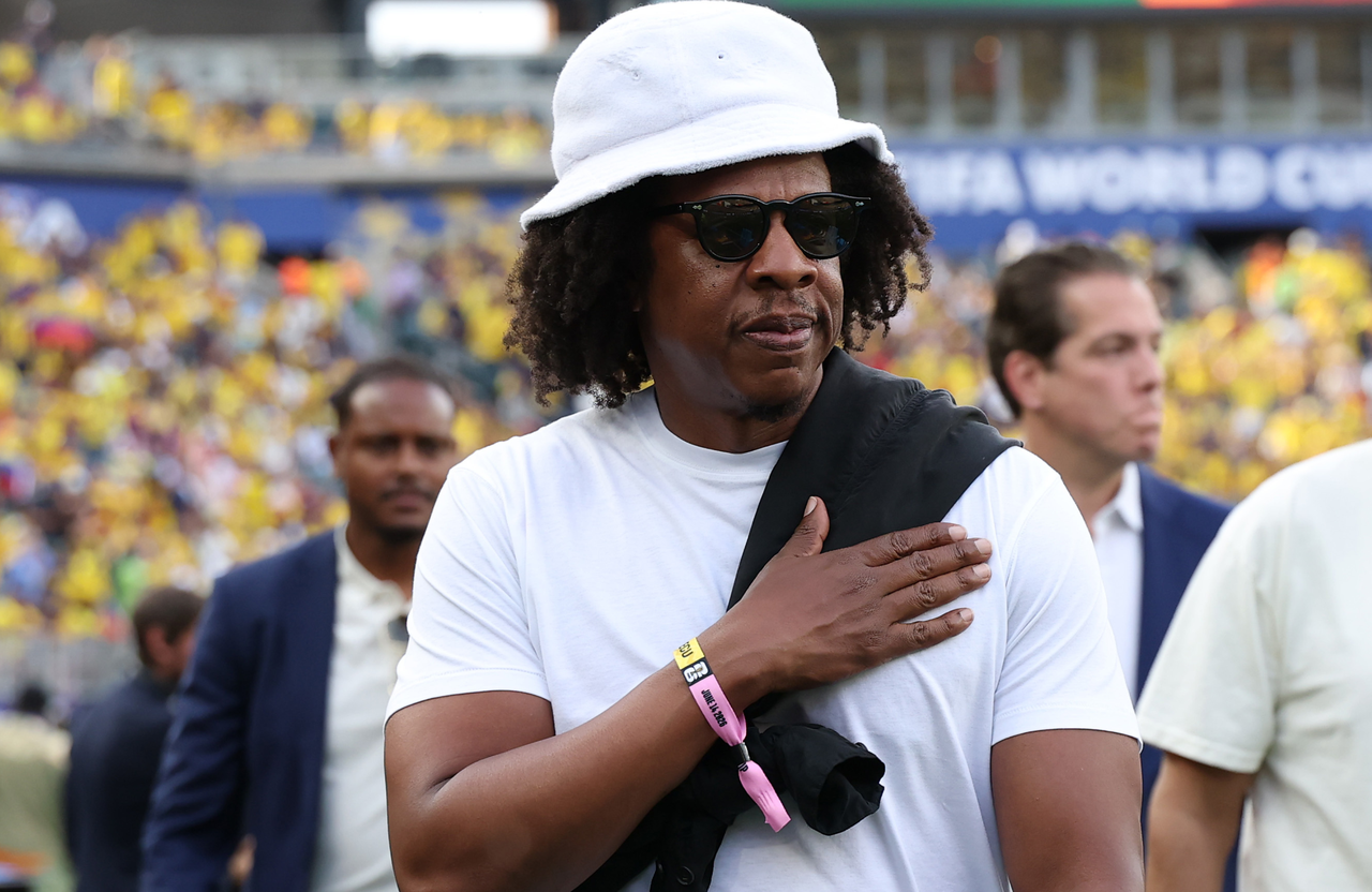 Jay-Z at a sports event, wearing a white bucket hat, sunglasses, and a white t-shirt. He has a hand on his chest.