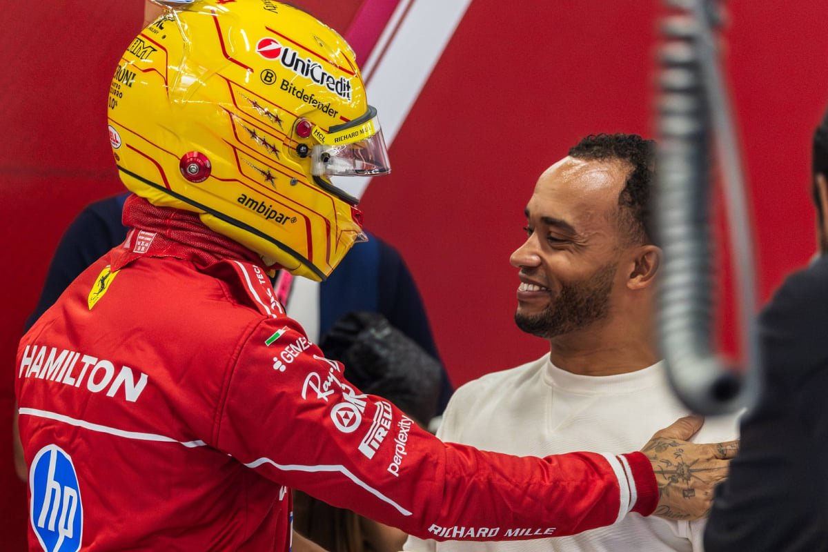 Lewis Hamilton in yellow helmet and red Ferrari care suit with a hand on the shoulder of brother Nicolas who is smiling in Ferrari garage — Photo: © IMAGO