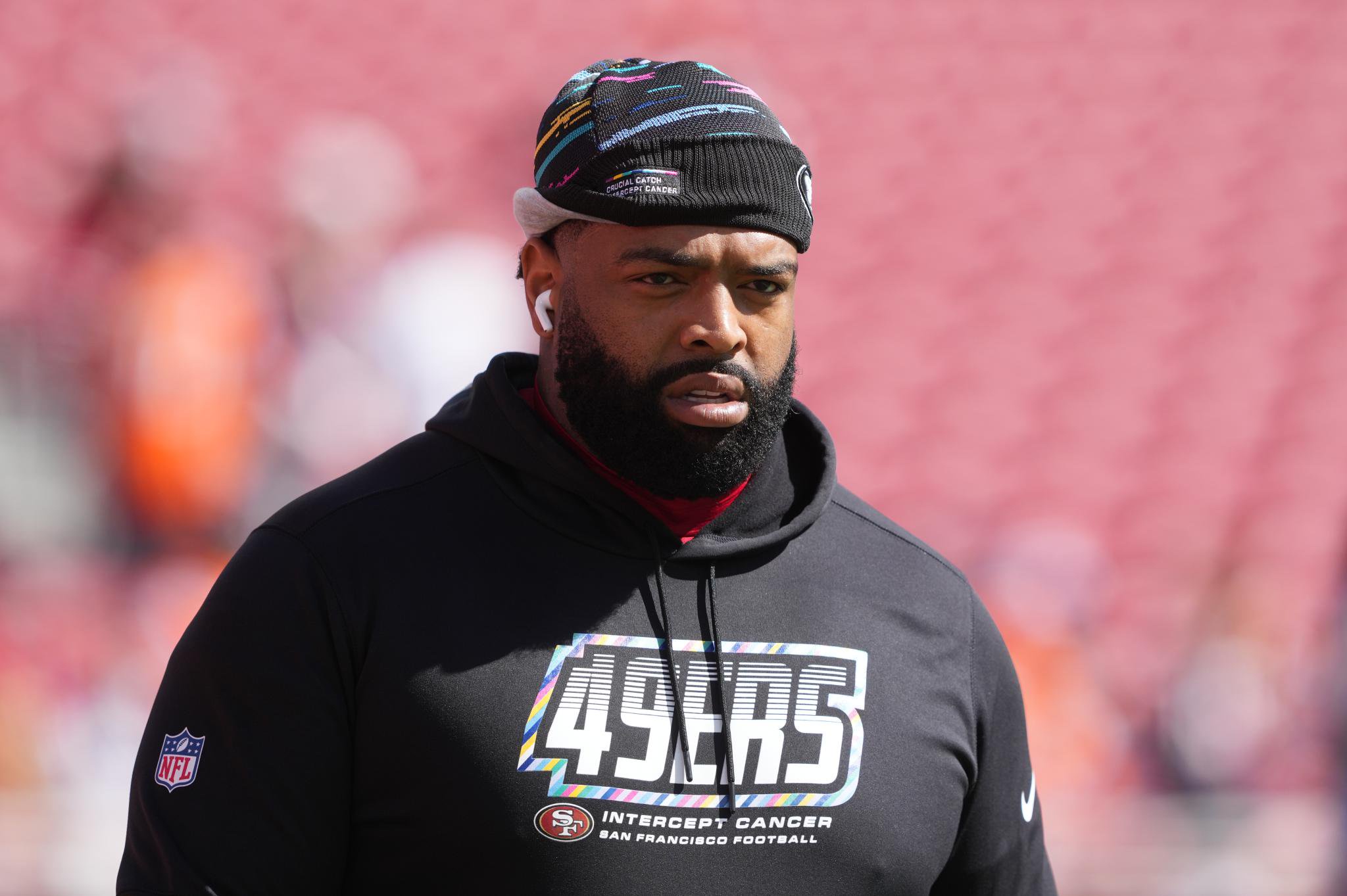San Francisco 49ers player wearing a black hoodie with "49ers" and "Intercept Cancer" text, and a matching patterned beanie.