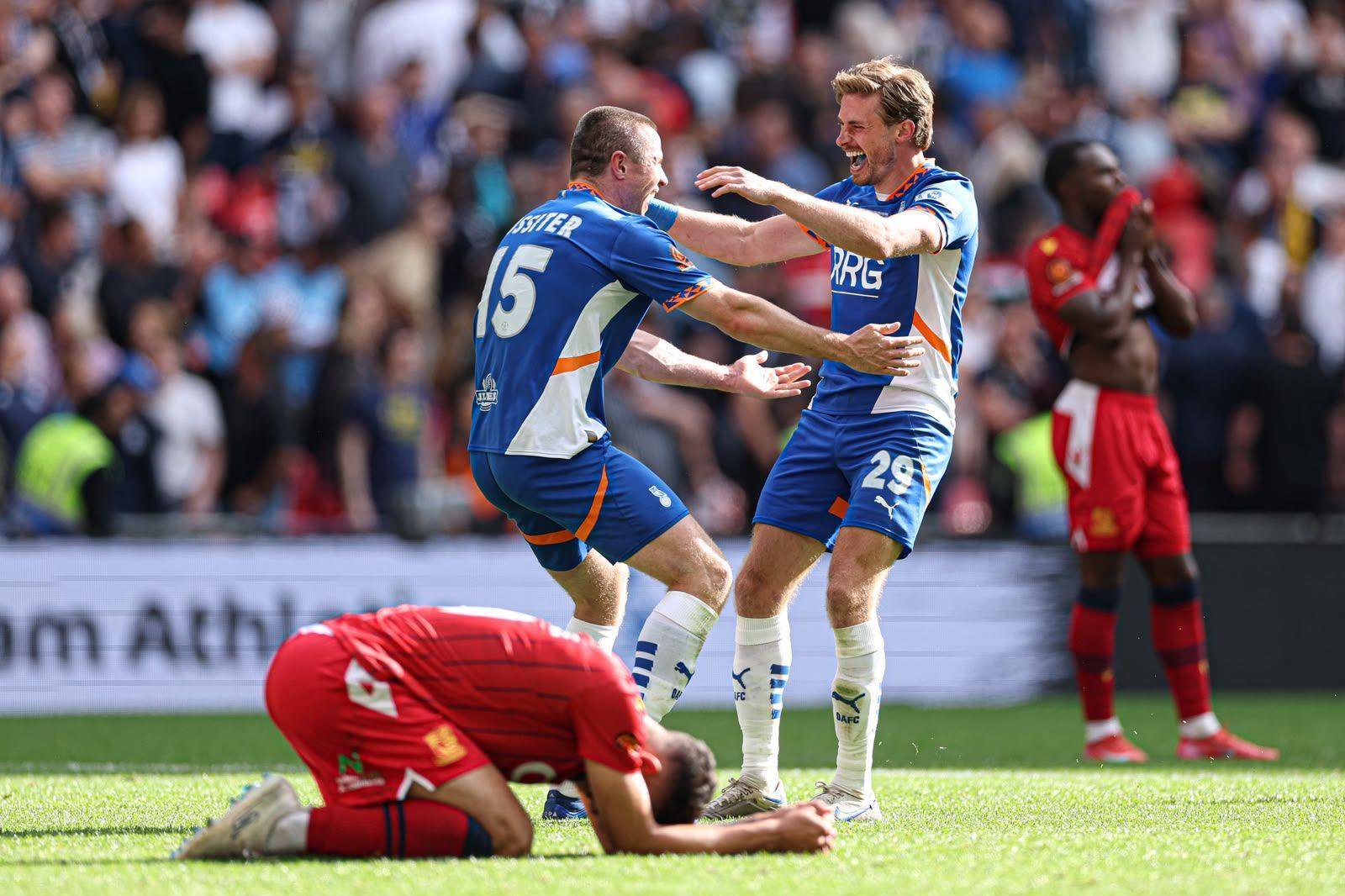 Two footballers in blue go to hug each other on a football pitch. Another player in red is crouched down on the turf, head in hands. Crowds on a stadium terrace are out of focus in the background.