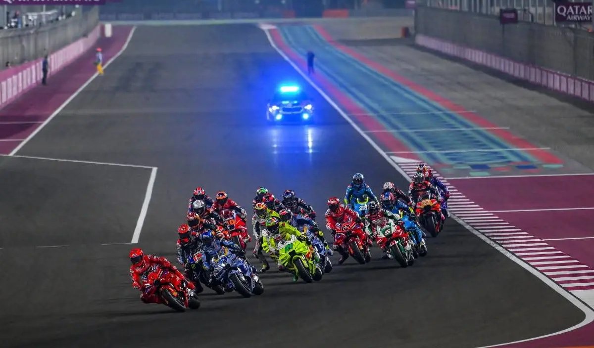 MotoGP riders lead a pack of bikes at night on the Losail circuit in Qatar, with a safety car ahead on track.