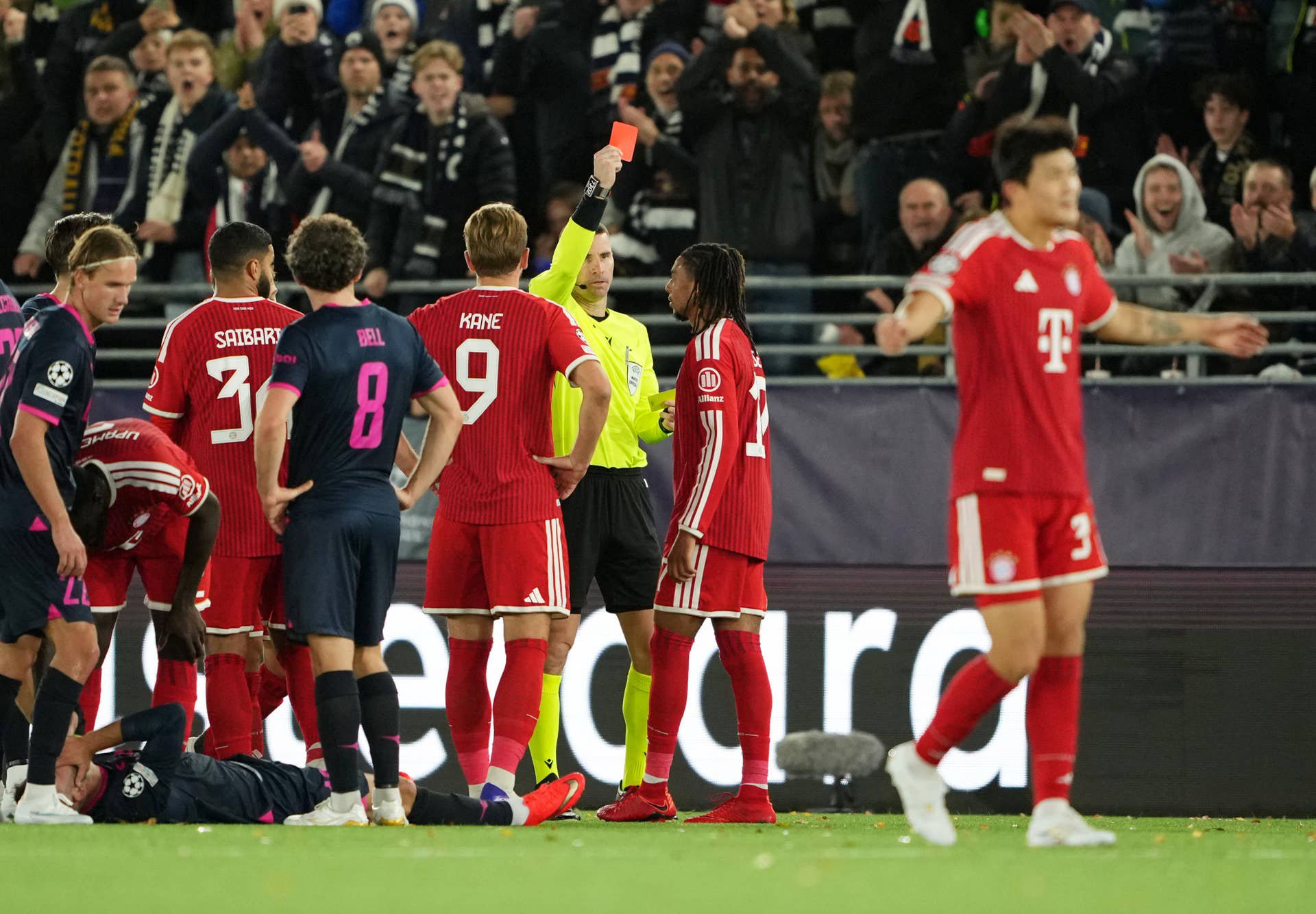 Ein Schiedsrichter zeigt einem Bayern-Spieler im Fußballspiel eine rote Karte, Spieler und Fans reagieren.