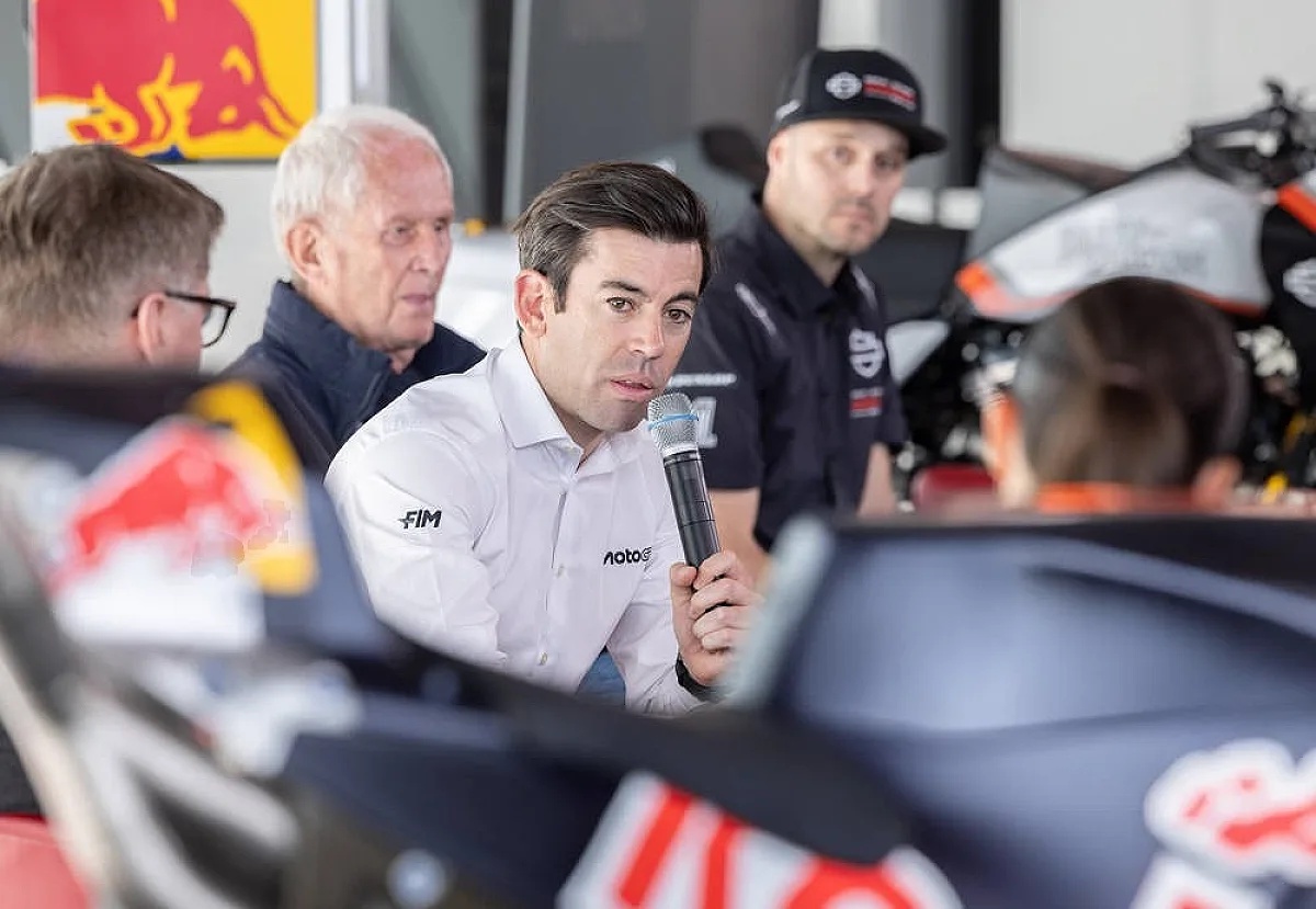 A man in a white FIM and Moto shirt speaks into a microphone at a MotoGP press event, with a Red Bull backdrop and a KTM bike behind him.