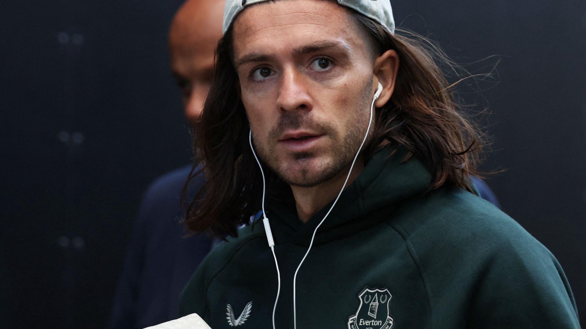 A man  wears a cap and has dark flowing long hair. He wears wired headphone and a tracksuit top with the Everton badge on it. 