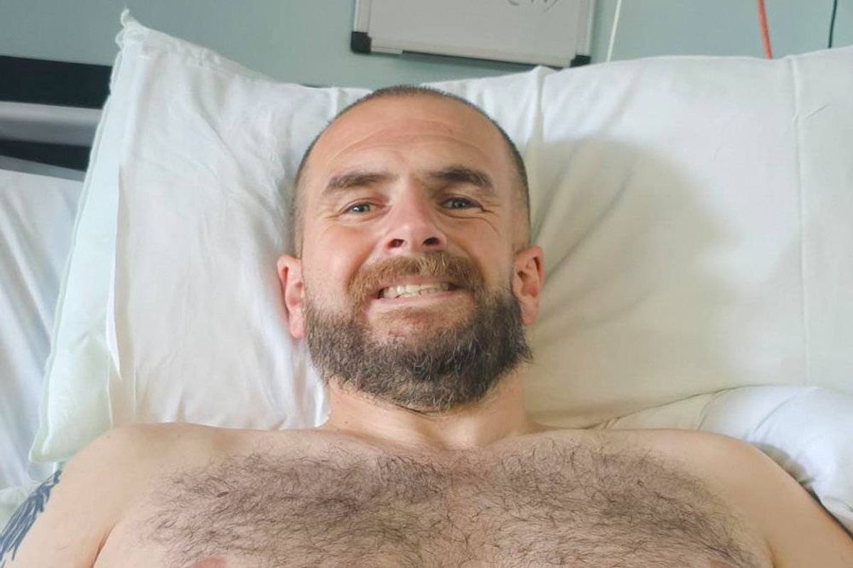 An individual, possibly a patient, reclines on a hospital bed, displaying a cheerful expression while surrounded by white pillows.