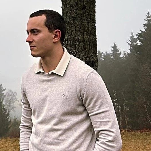 Ein junger Mann mit kurzen dunklen Haaren, der einen hellbeigen Pullover über einem cremefarbenen Hemd mit Kragen trägt, steht im Freien vor einem Baum. Im Hintergrund ist eine neblige Waldlandschaft mit Bäumen und braunem Gras zu sehen. Er blickt nachdenklich nach links. Diese Beschreibung wurde mit der FootballR KI automatisch generiert.