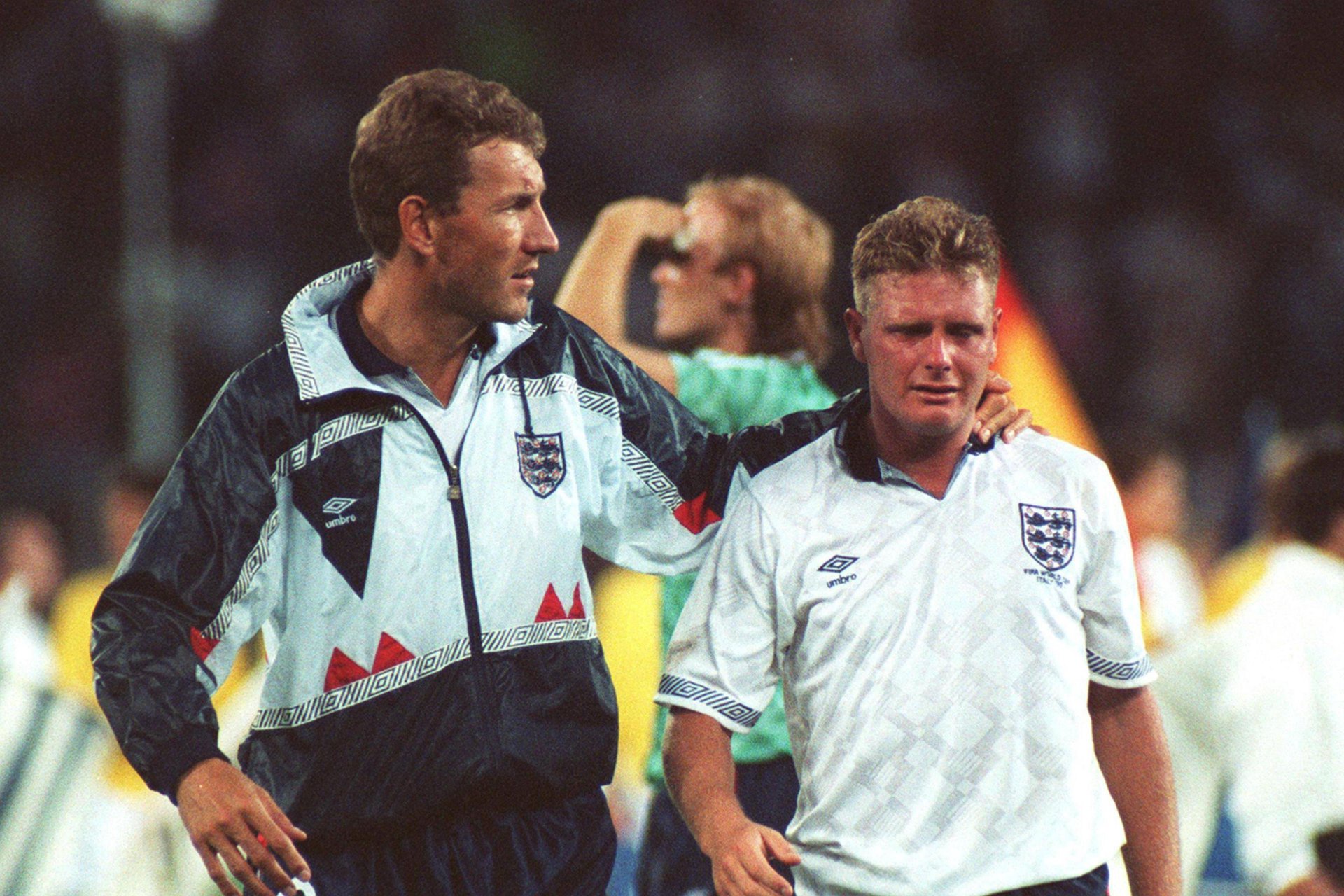 Terry Butcher in football kit and a football training jacket, walks on a football pitch, towards the camera, with his arm around the neck of Paul Gascoigne, who is on his left. Gascoigne wears a white England shirt, with the Three Lions' crest, and he is crying.