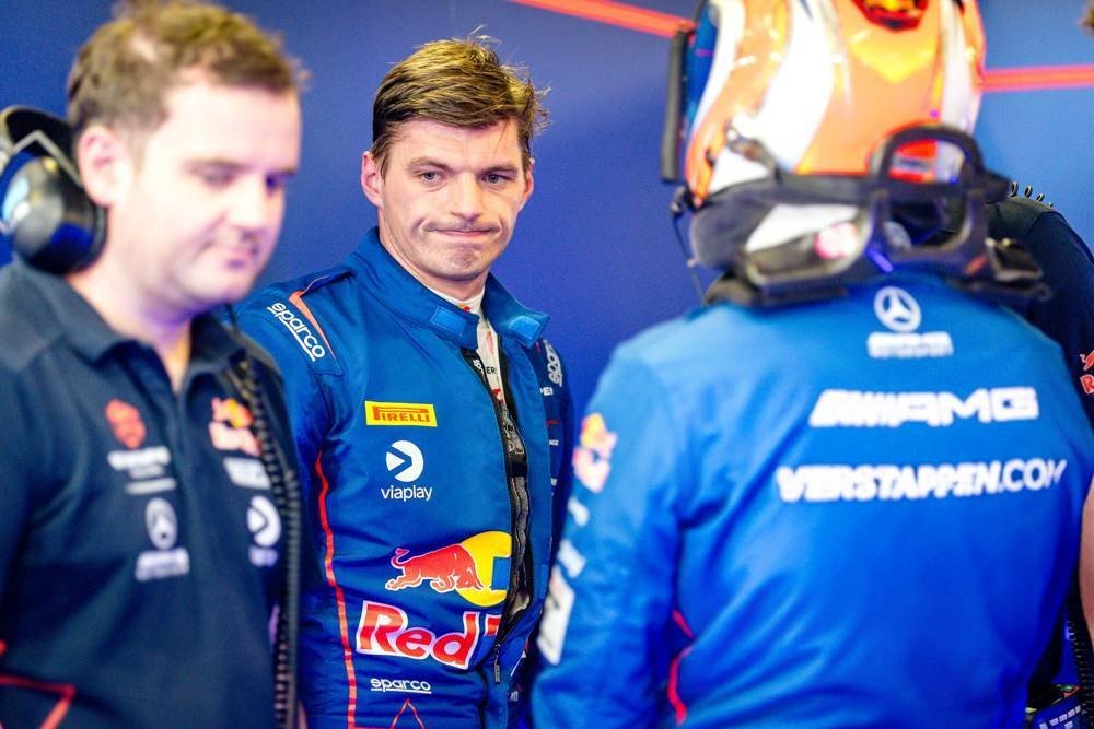 Max Verstappen in a blue Red Bull racing suit, with Verstappen.com and AMG Mercedes branding on a teammate's suit, in the F1 garage.