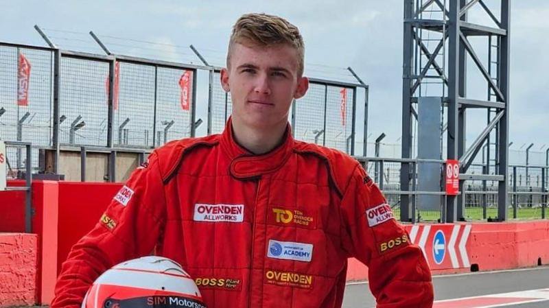 A young, blonde teenage boy in a red race suit. He has a red and white helmet under his arm.