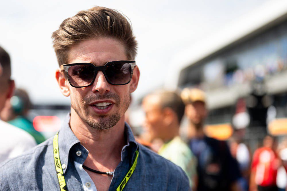 A man with swept-back hair, dark sunglasses and a stubbled beard wears a blue linen shirt and a yellow lanyard at a busy race paddock.
