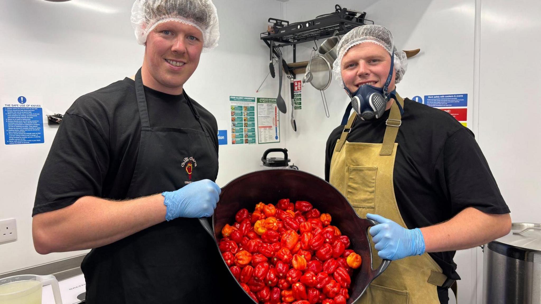 Two young men in hairnets and gloves and black t-shirts hold up a big bucket of red chillies