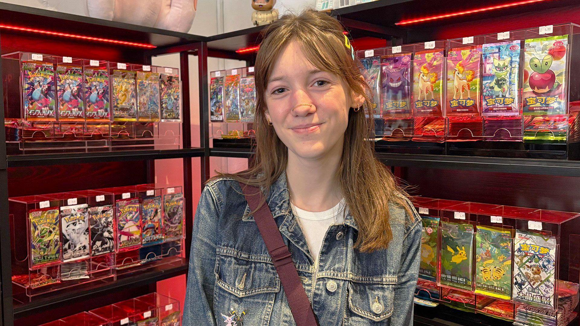 A young woman with dark hair, wearing a denim jacket with a Pokémon badge on it is smiling at the camera. She is standing in front of a display of Pokémon cards in a shop.