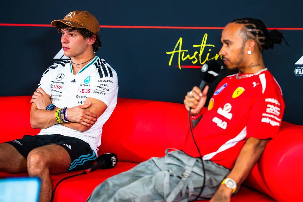 Two drivers in F1 team gear sit on a red sofa at Austin; one wears a Mercedes cap and white shirt, the other a red Ferrari shirt and holds a microphone.