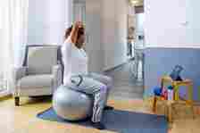 Senior woman doing stretching exercise on fitness ball while watching online fitness tutorial.