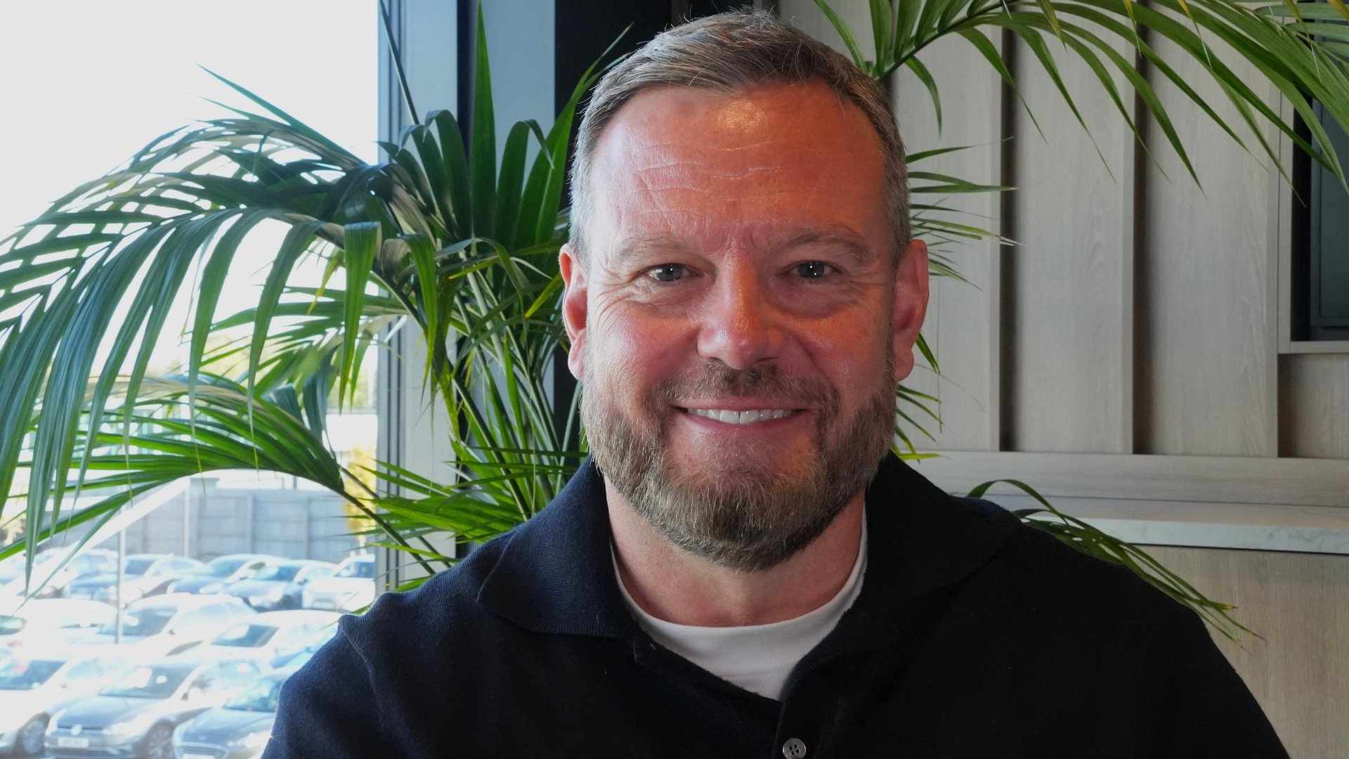 Mark Ashton smiling directly at the camera. He is in an office with a potted plant behind him. He has short hair and a beard and is wearing a navy top over a white T-shirt.