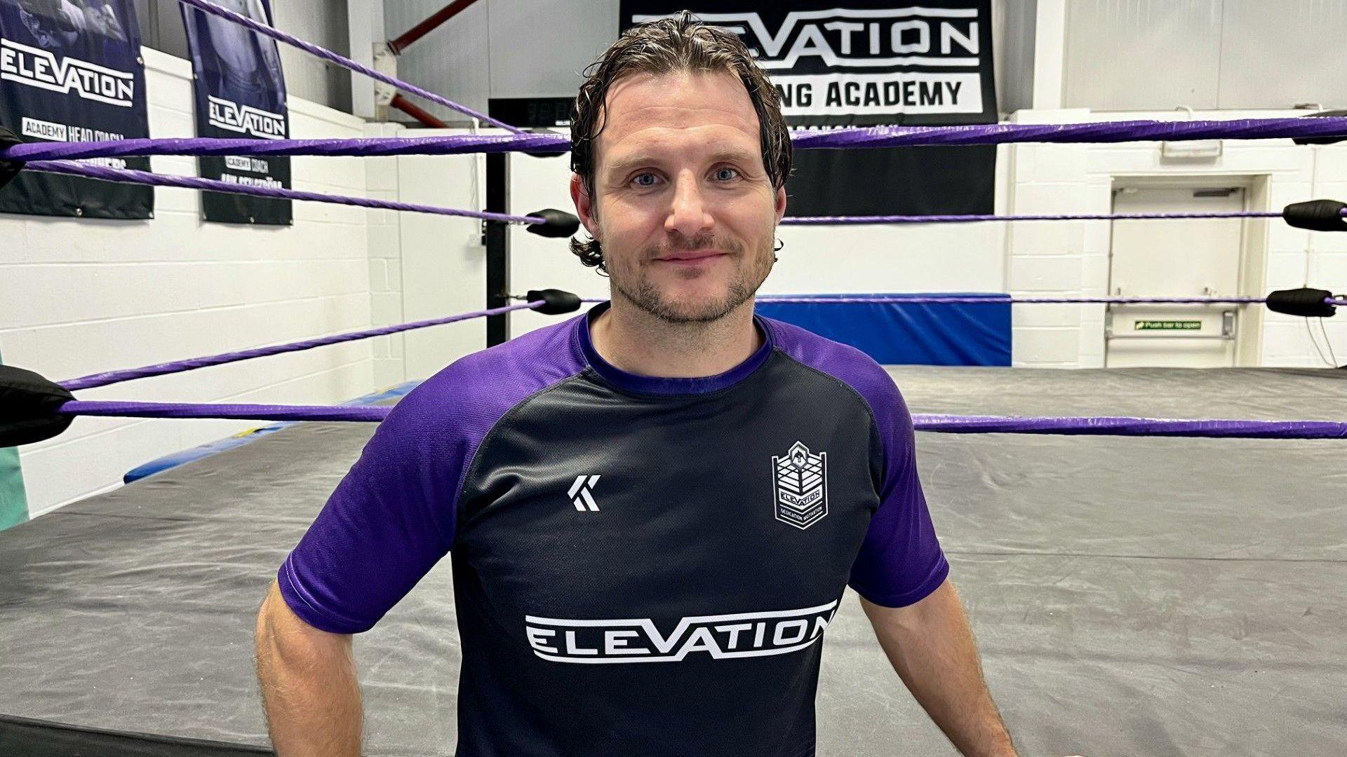 A man smiles for the camera. He has short dark hair and is wearing a T-shirt with the word "Elevation" on the front. Behind him is a wrestling ring.