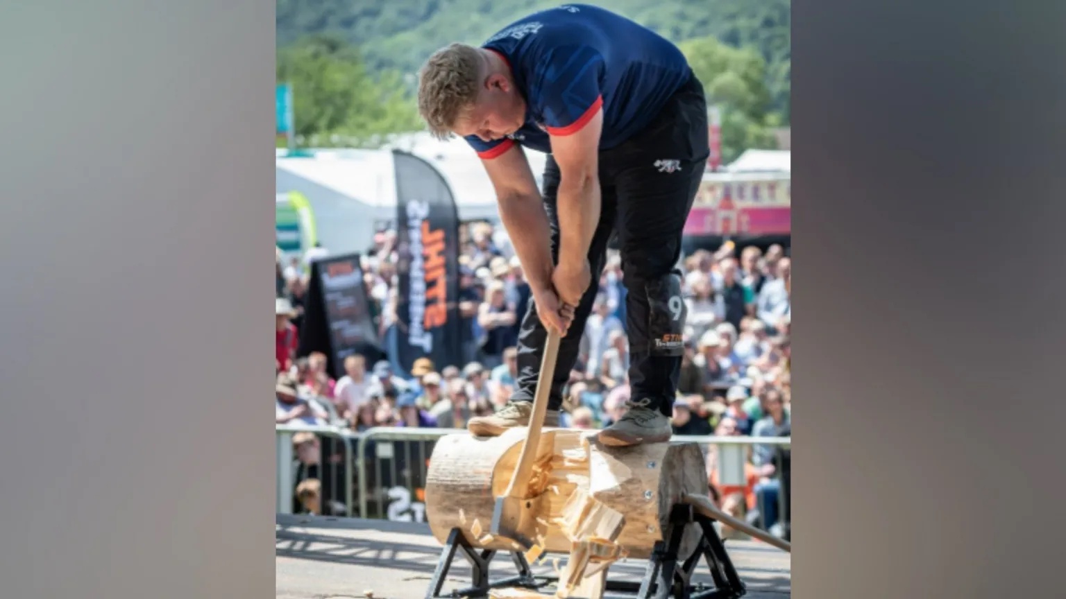 STIHL Timbersports Spencer stands on a small log and swings at it with an axe. A large crowd of spectators are seen behind him.