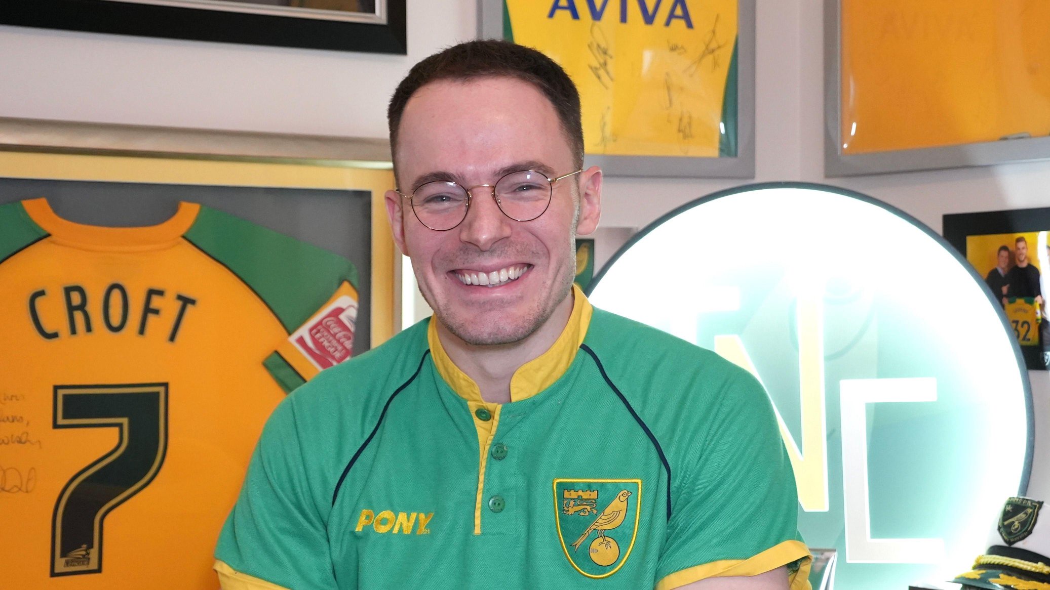 Chris Reeve in his Talk Norwich City studio. He is smiling and wearing a green Norwich shirt with other commemorative yellow shirts in picture frames dotted on the wall behind. One says "Croft 7" on the back.