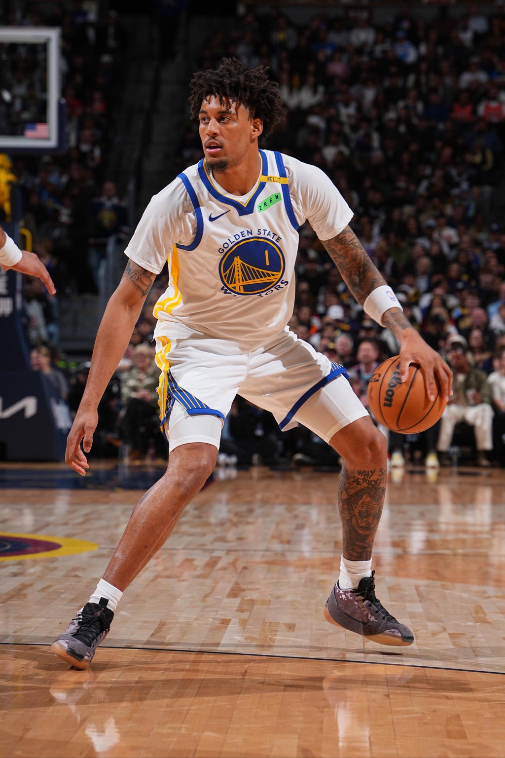 A basketball player wearing a white Golden State Warriors jersey dribbles the ball on the court.