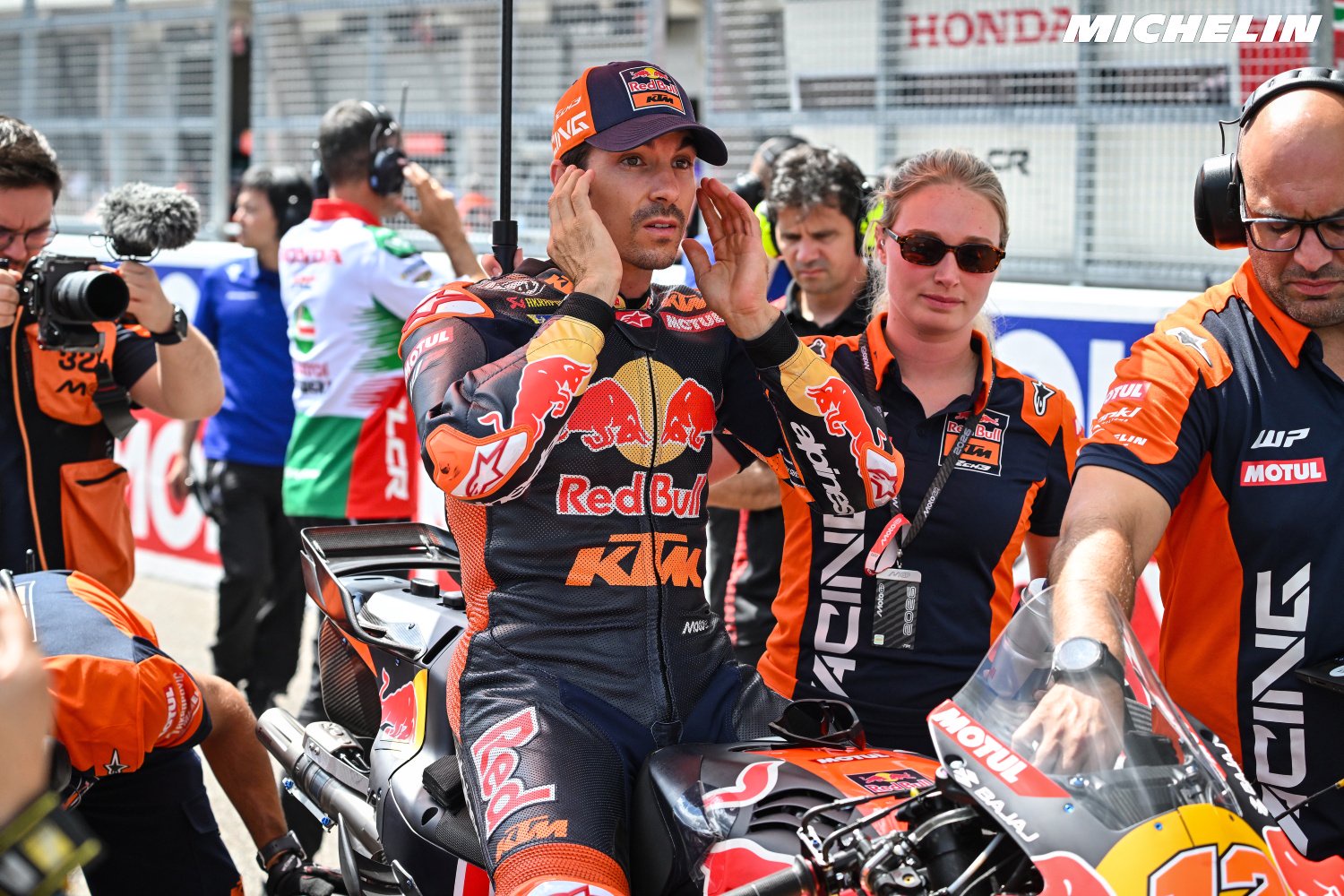 Maverick Viñales in Red Bull KTM racing suit and cap, hands on his helmet at the MotoGP paddock beside his KTM bike, surrounded by team crew and photographers