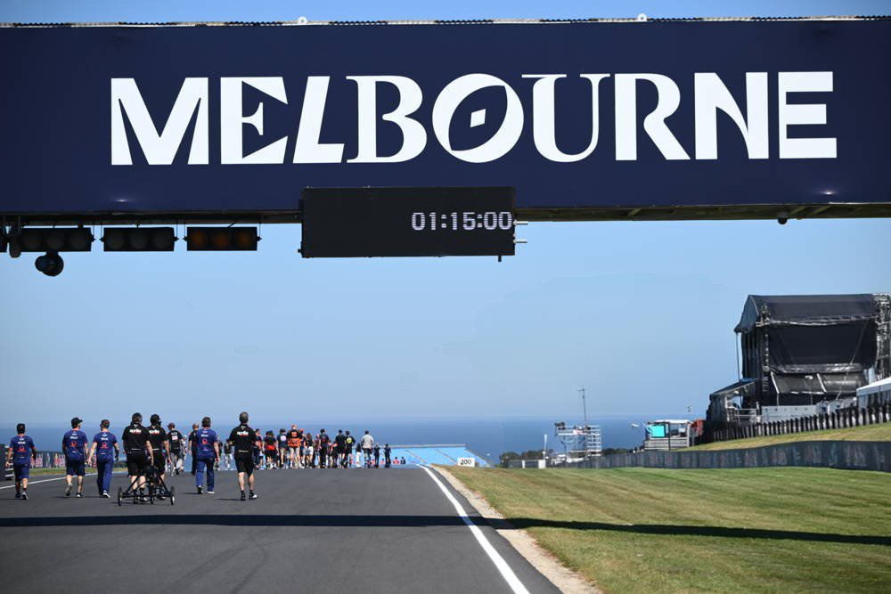 Marshals and staff in blue and black uniforms walk along the Phillip Island circuit straight under a "MELBOURNE" banner showing 01:15:00 on the timing board.
