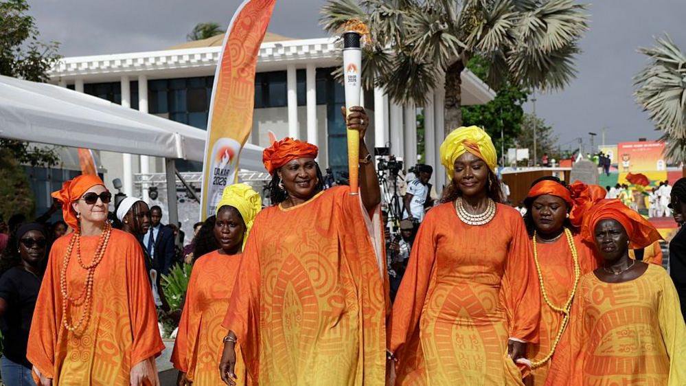 A group of women in matching long orange-and-yellow gradient traditional gowns walk in procession as the central woman proudly holds up a lit Dakar 2026 Youth Olympic Games torch outdoors. All of the women wear headscarfs which are either orange or yellow