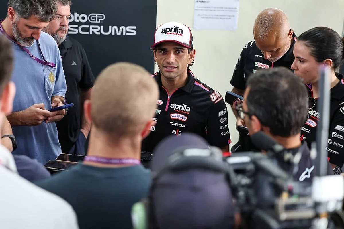 A rider in an Aprilia cap and black Aprilia race suit speaks to reporters in the MotoGP paddock, with a cameraman in the foreground.