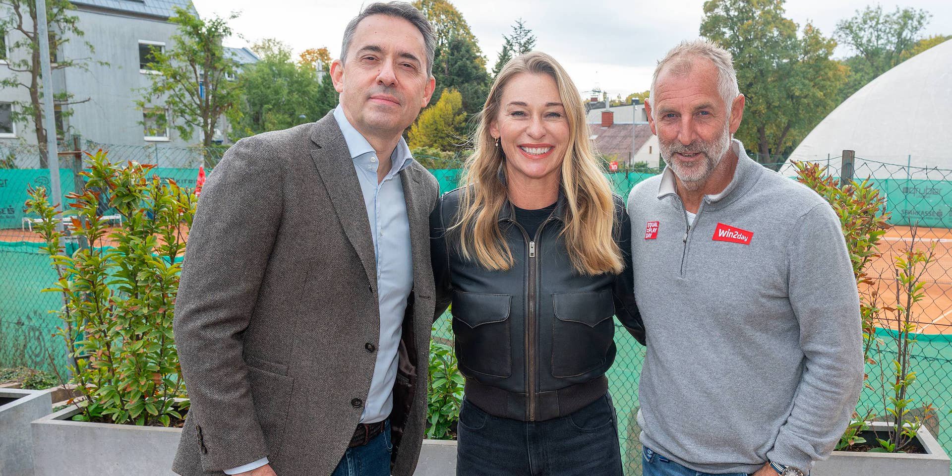 Drei Personen posieren lächelnd vor einem Tennisplatz im Freien.