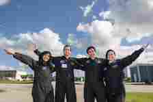 Four NASA personnel in black jumpsuits stand outside and smile with their arms outstretched. The background features a bright blue sky with scattered clouds and some buildings.