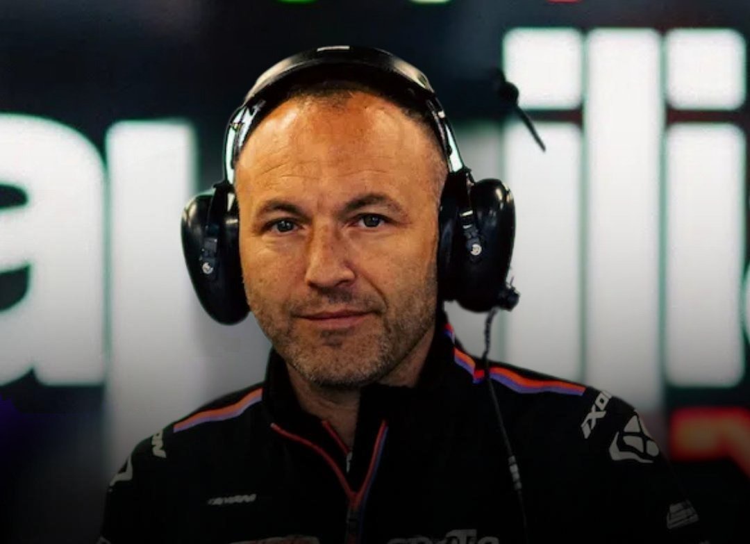 Man with headset and black team jacket in a pit garage, background with blurred white lettering; likely a MotoGP team official