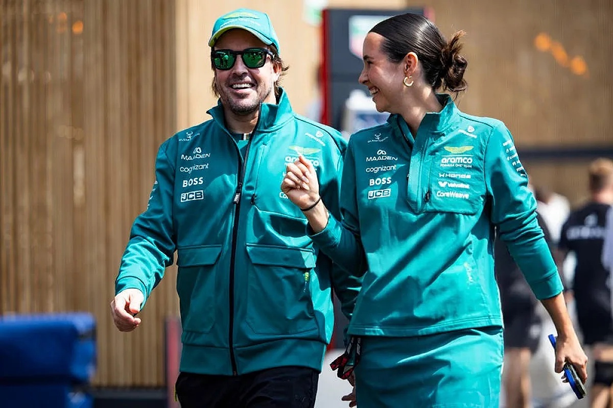 Two people in teal Aston Martin F1 team jackets laughing in the paddock, one wearing a green cap and sunglasses.
