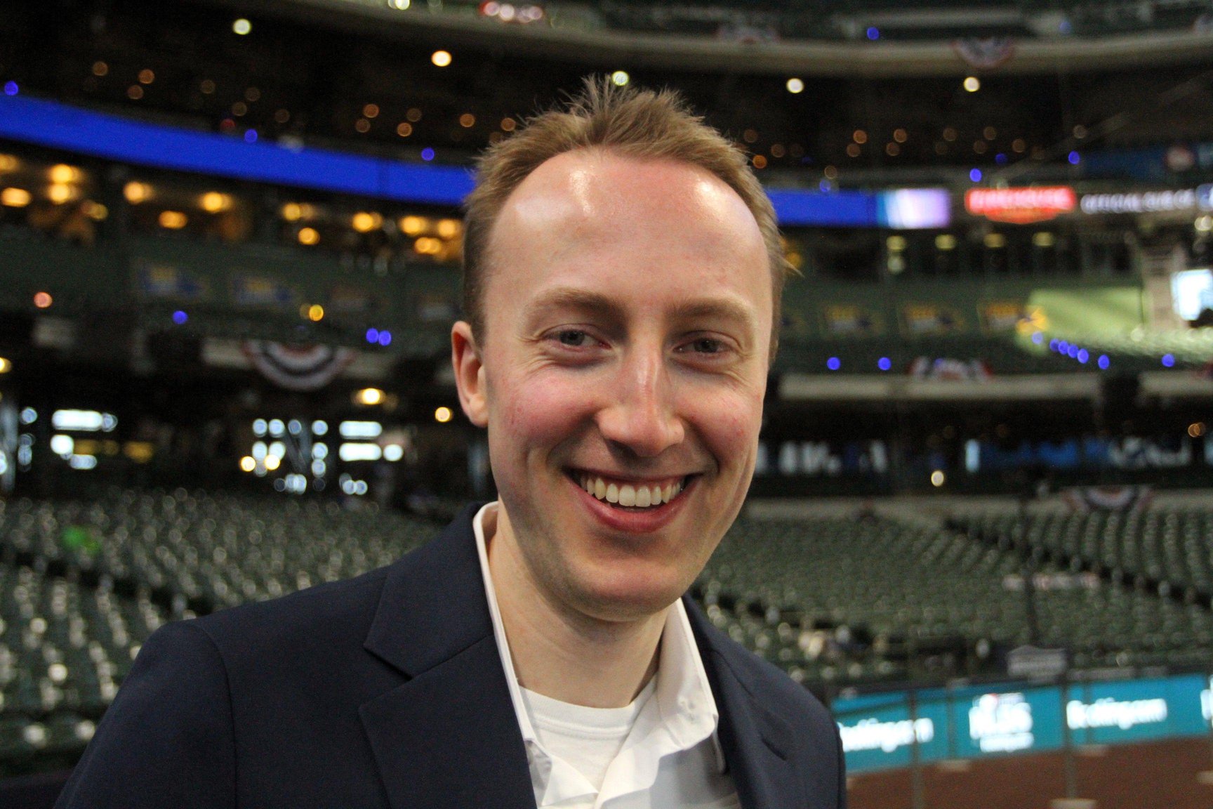 Hunter Baumgardt is pictured inside a baseball stadium in America. He is wearing a navy blue overcoat and white shirt. There are thousands of empty seats behind him.