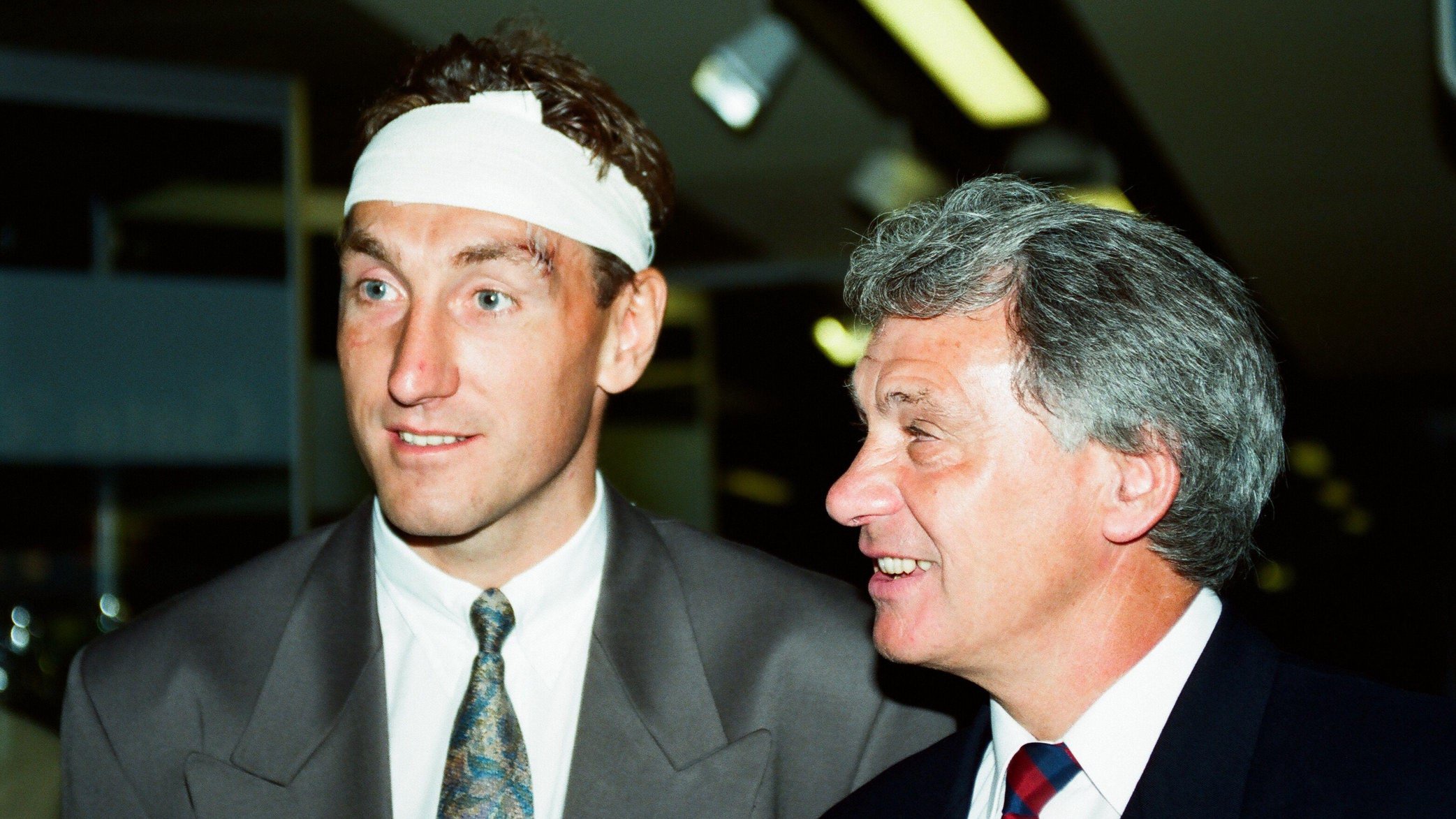 Terry Butcher on the left is wearing a suit. He has a bandage around his head and a wound on his eyebrow. Sir Bobby Robson is wearing a suit and tie. They are both smiling.