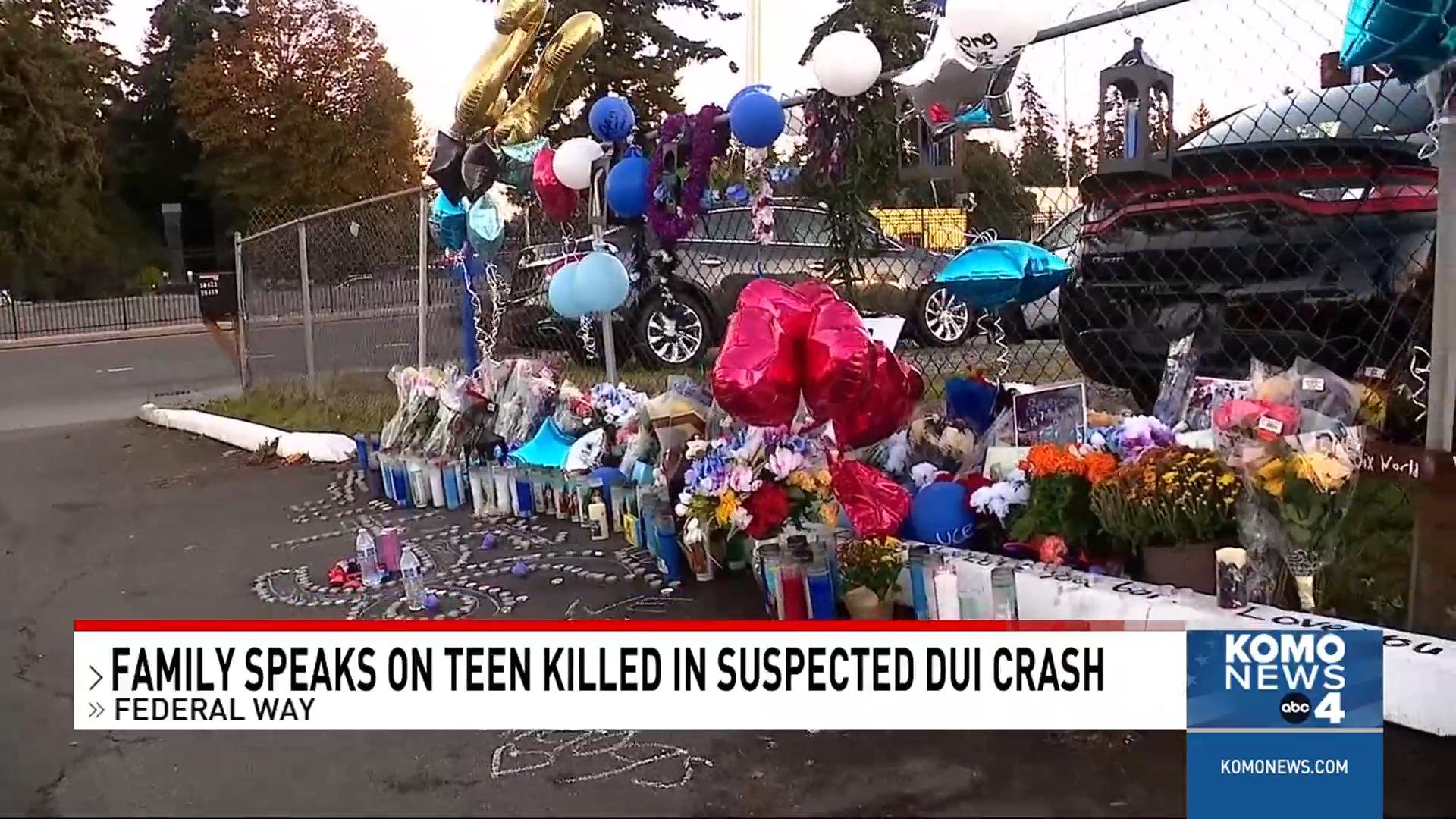 A roadside memorial with flowers, balloons, candles, and framed pictures along a chain-link fence, with a news chyron at the bottom.