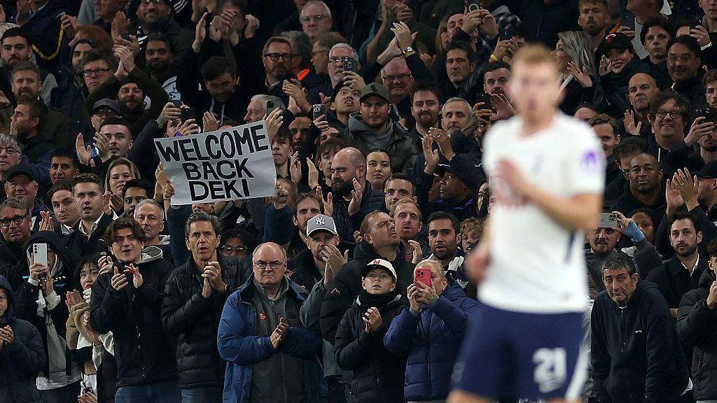 A fan in the crowd holds a sign saying "welcome back Deki". Dejan Kulusevski is out of focus in the foreground.