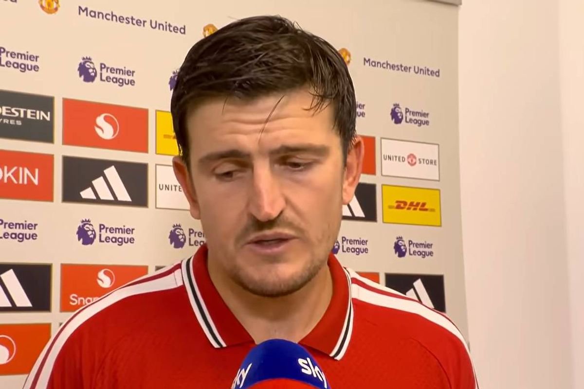 An individual dressed in a red polo shirt stands before a microphone, addressing a gathering in a press conference setting. Behind him, a backdrop adorned with various logos and branding elements can be observed.
