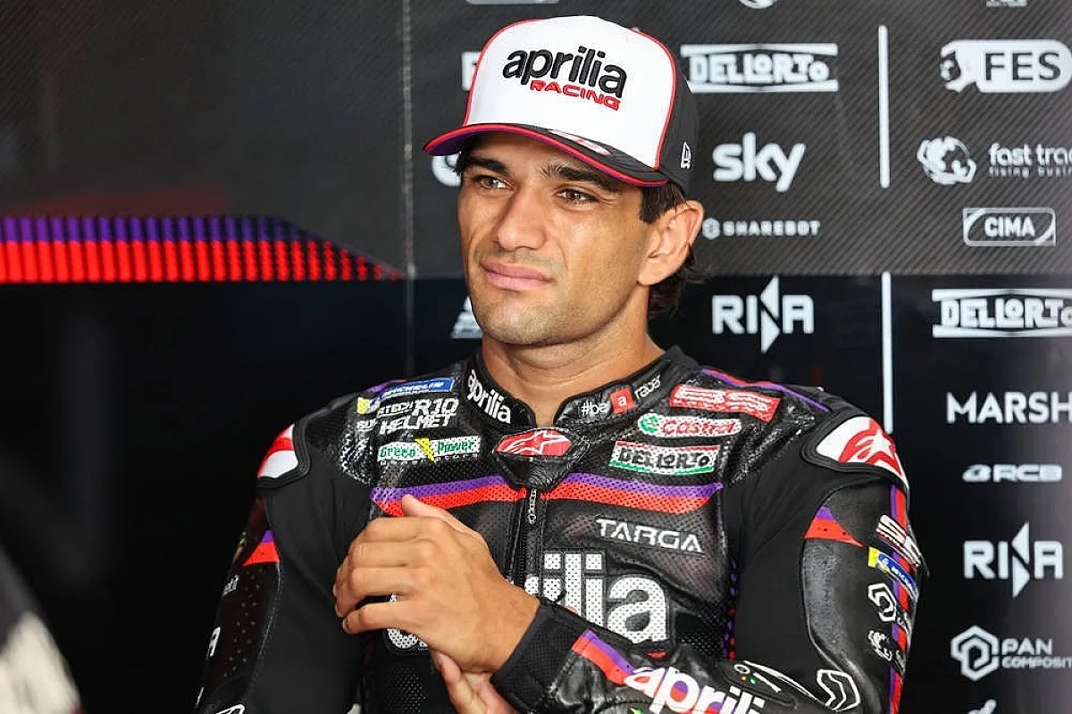 A rider in a black Aprilia Racing suit and white cap in the MotoGP paddock, with Aprilia, Castrol and Sky logos behind him.