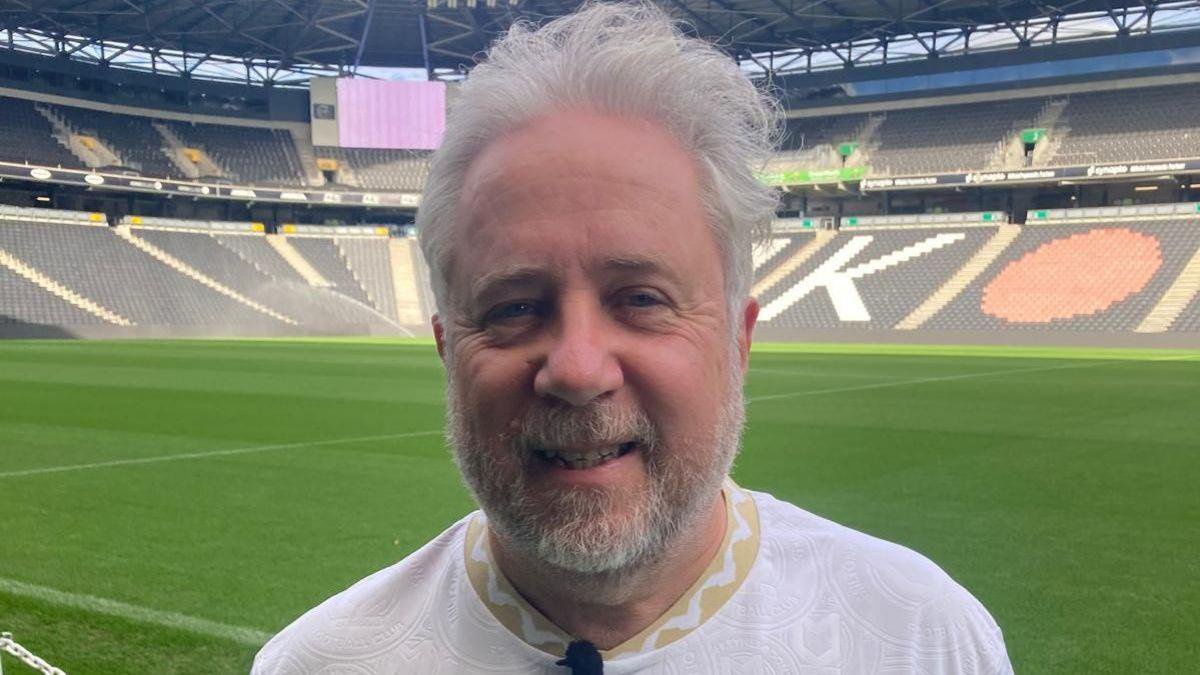MK Dons fan Jon Scudamore is standing beside a green football pitch at Stadium MK wearing a white shirt. 