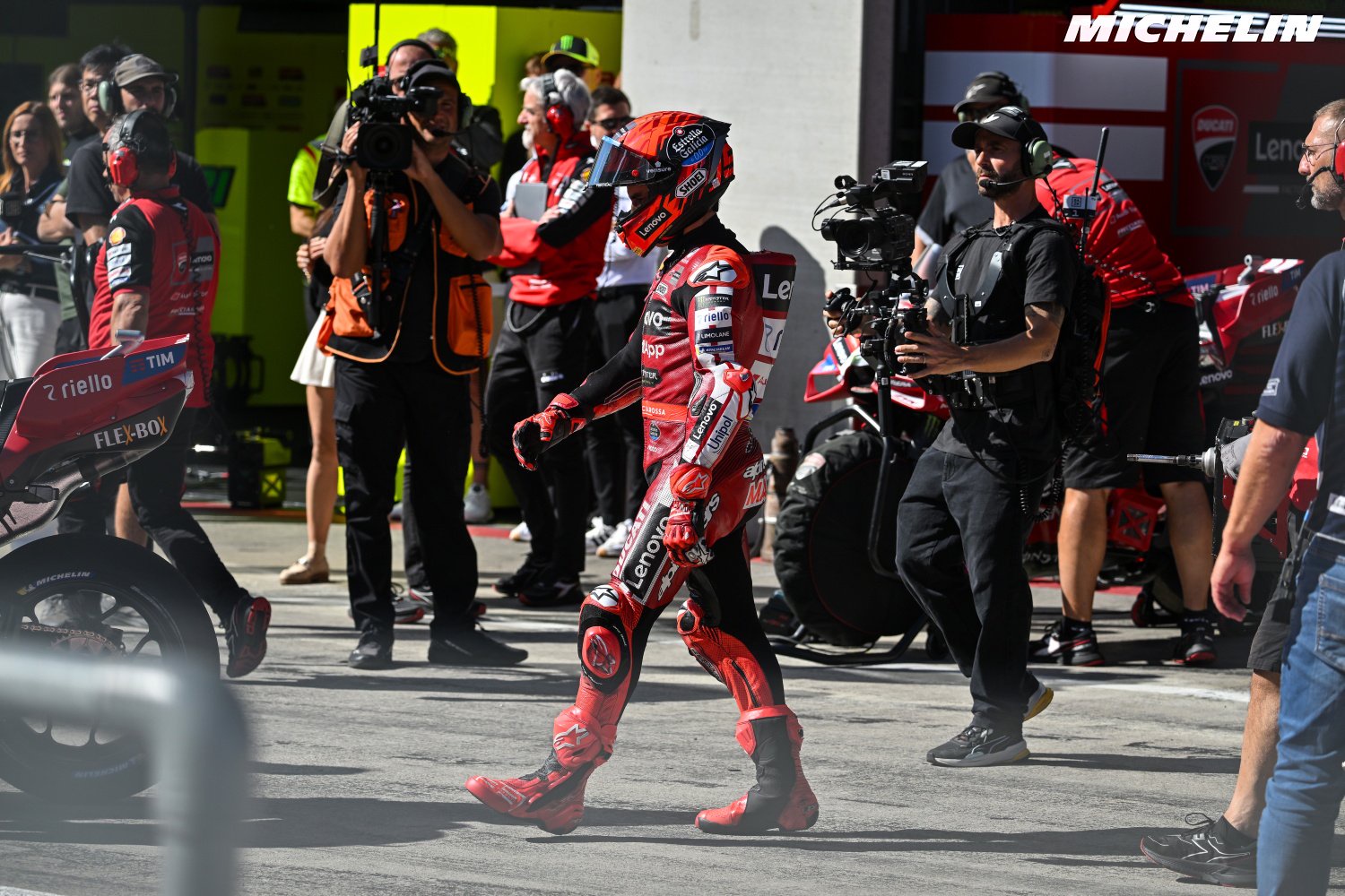 A MotoGP rider in red Ducati leathers walks through the pit lane past cameras and crew, with a Ducati bike beside him.