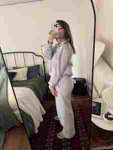 A person in gray loungewear takes a mirror selfie in a bedroom with a dark green bedspread and a red patterned rug.