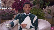 A man sitting in a talk show environment, wearing a green and white sweater with a diagonal pattern