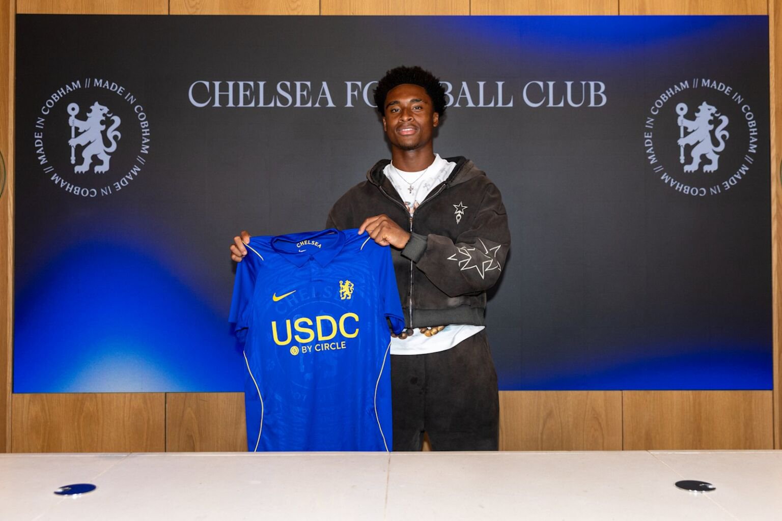 Chizaram Ezenwata holding a Chelsea shirt in front of a Made in Cobham backdrop after signing his new contract