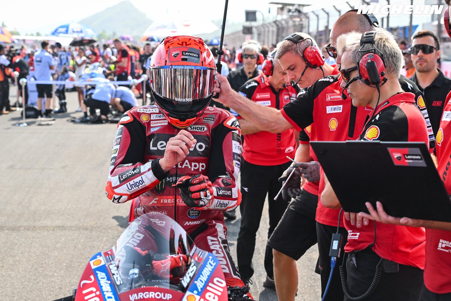 Rider in red Ducati leathers and helmet on the grid at MotoGP, with Ducati crew in red beside him, one holding a tablet