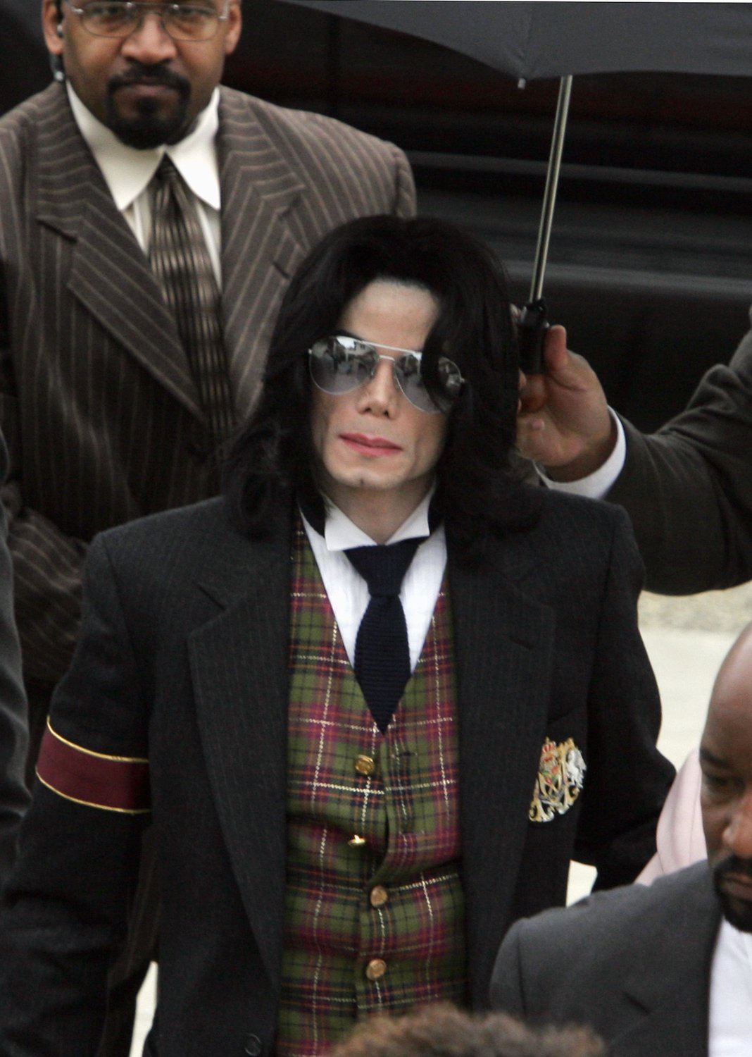 Michael Jackson wearing a dark suit with a plaid vest, aviator sunglasses, and dark hair, surrounded by security, as he arrives for his child molestation trial.