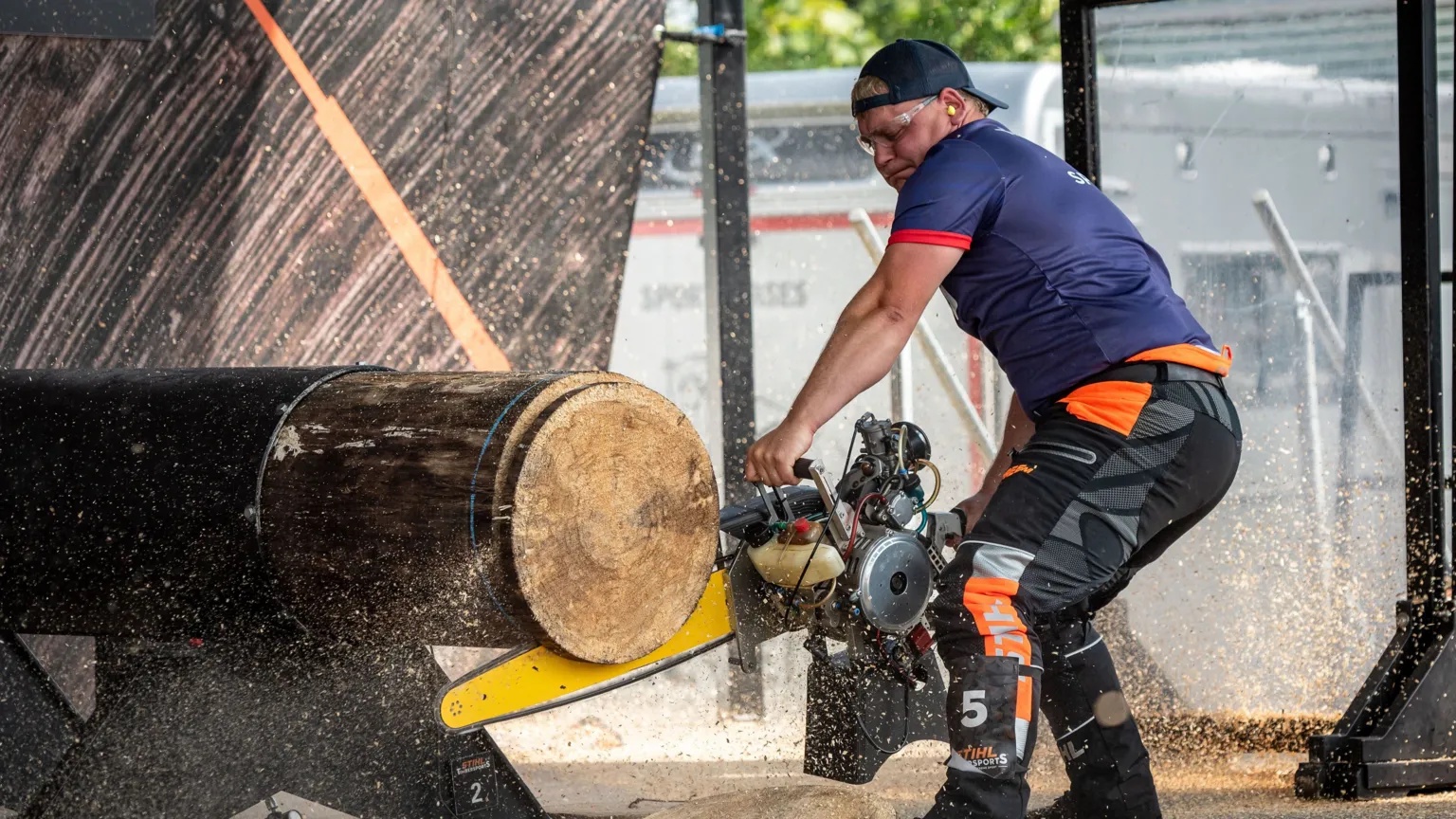 STIHL Timbersports George Spencer uses a chainsaw to cut through a large log. He is wearing a navy blue top, black cap and black and orange trousers. 