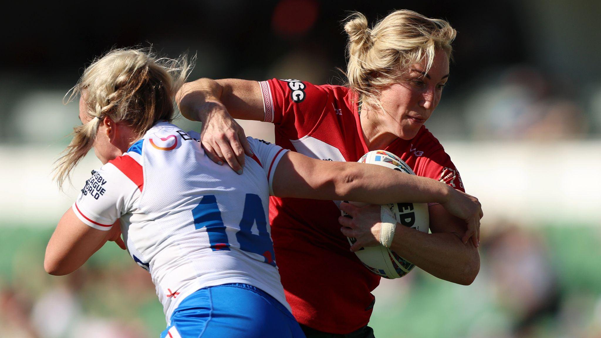 Bethan Dainton in the red of Wales runs with the ball as she tries to evade a tackle from a white-shirted England player.