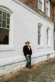 A person wearing a cowboy hat, dark blazer, jeans, and brown shoes leans against a white brick building with three arched windows. The ground has patches of grass. The person is looking at the camera with one hand in their pocket.