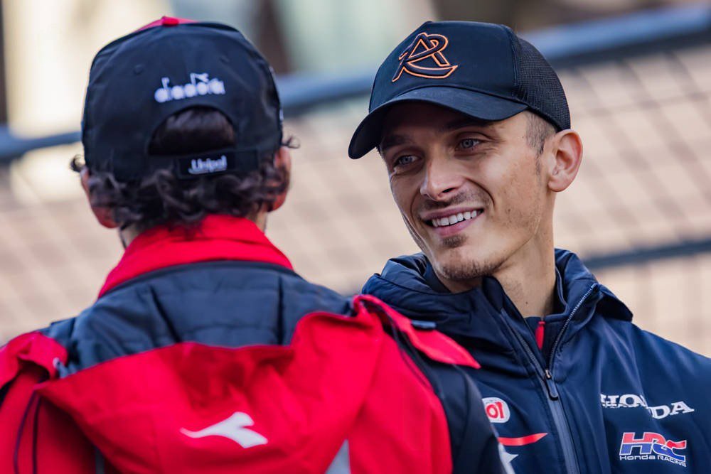 Two men in MotoGP team gear in a paddock: one in a black cap and Honda HRC jacket smiling, the other seen from behind in a red Diadora jacket.