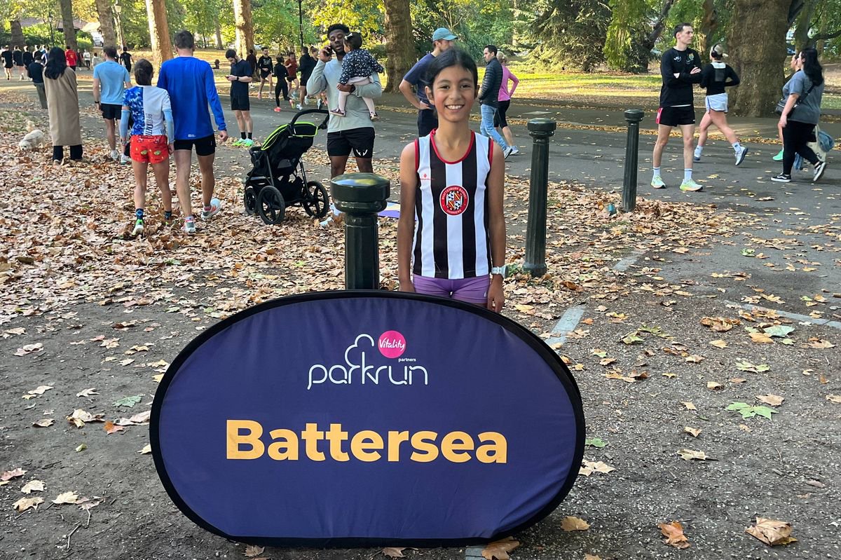 An individual standing next to a large rectangular sign on a path, surrounded by an outdoor setting with numerous individuals walking. The sign displays the word "Battersea" in bold, yellow letters against a blue background.